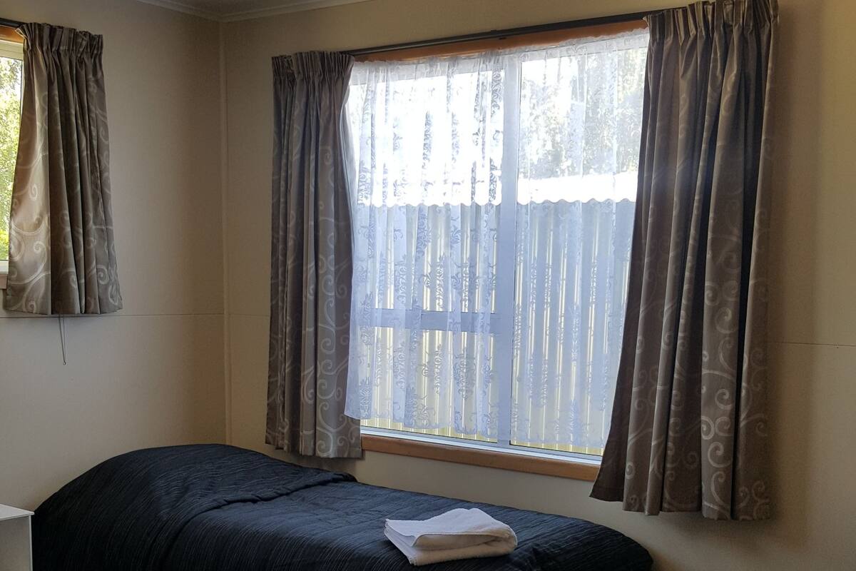 Ein Zimmer mit zwei Betten, einem Vorhangfenster, einem Deckenventilator und einer kleinen Ablagefläche mit Gegenständen.