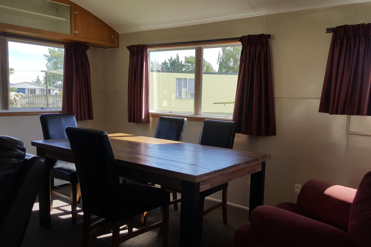 Ein Zimmer mit Holztisch und Stühlen, großen Fenstern mit Vorhängen und Blick auf ein Haus.