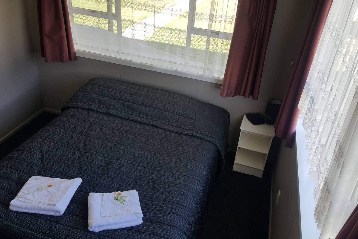 Ein kleines, ordentlich gemachtes Bett mit einem dunkelblauen Überwurf und zwei gefalteten weißen Handtüchern. Ein Fenster bietet Blick auf grüne Vegetation, und ein weißer Nachttisch mit Lampe und einem kleinen Gegenstand darauf.