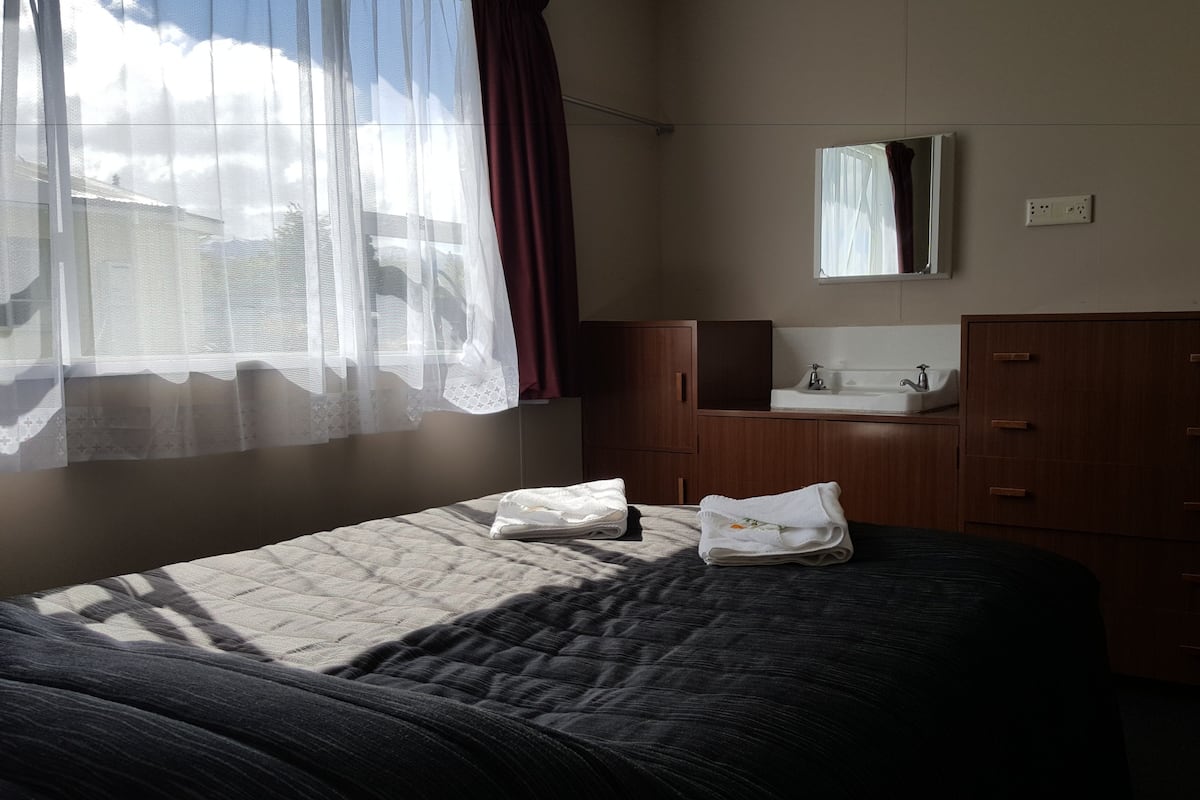 Ein Hotelzimmer mit Bett, Badezimmer mit Waschbecken und Spiegel sowie einem Fenster mit durchsichtigen Vorhängen.
