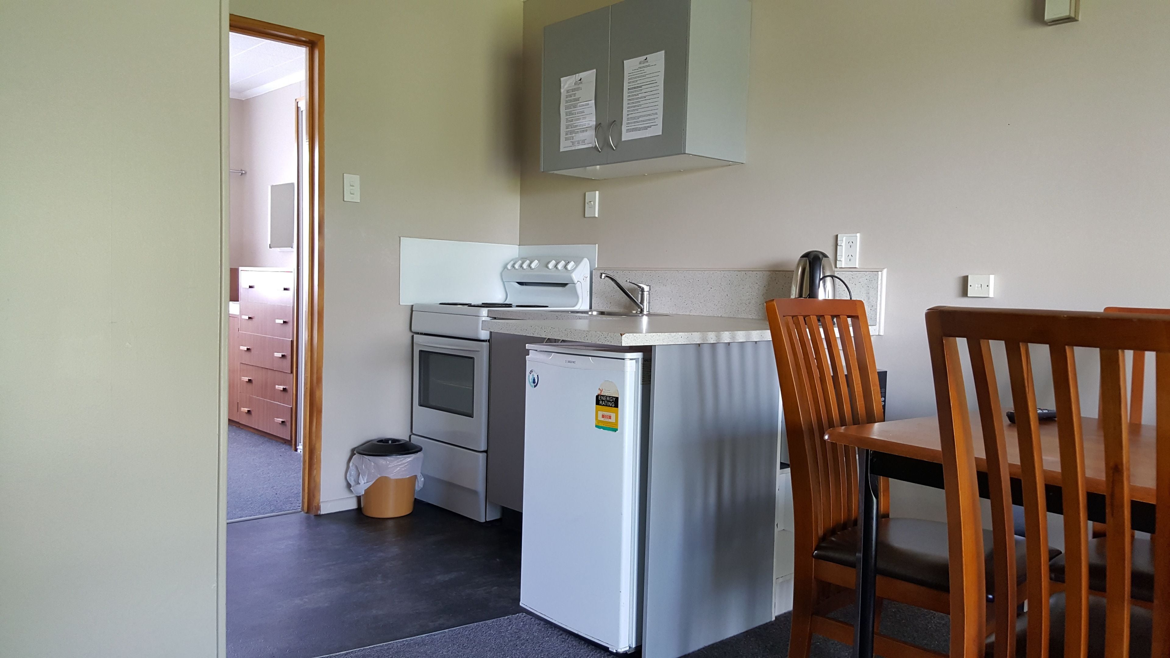 A compact kitchen with a refrigerator, oven, microwave, and a small sink. There is a wooden chair and a trash bin.