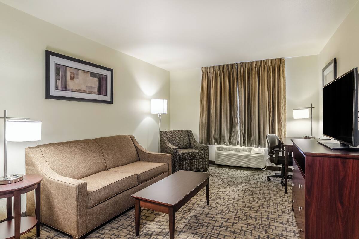 A hotel room with a sofa, armchair, TV, desk, lamp, air conditioning unit, and curtains.