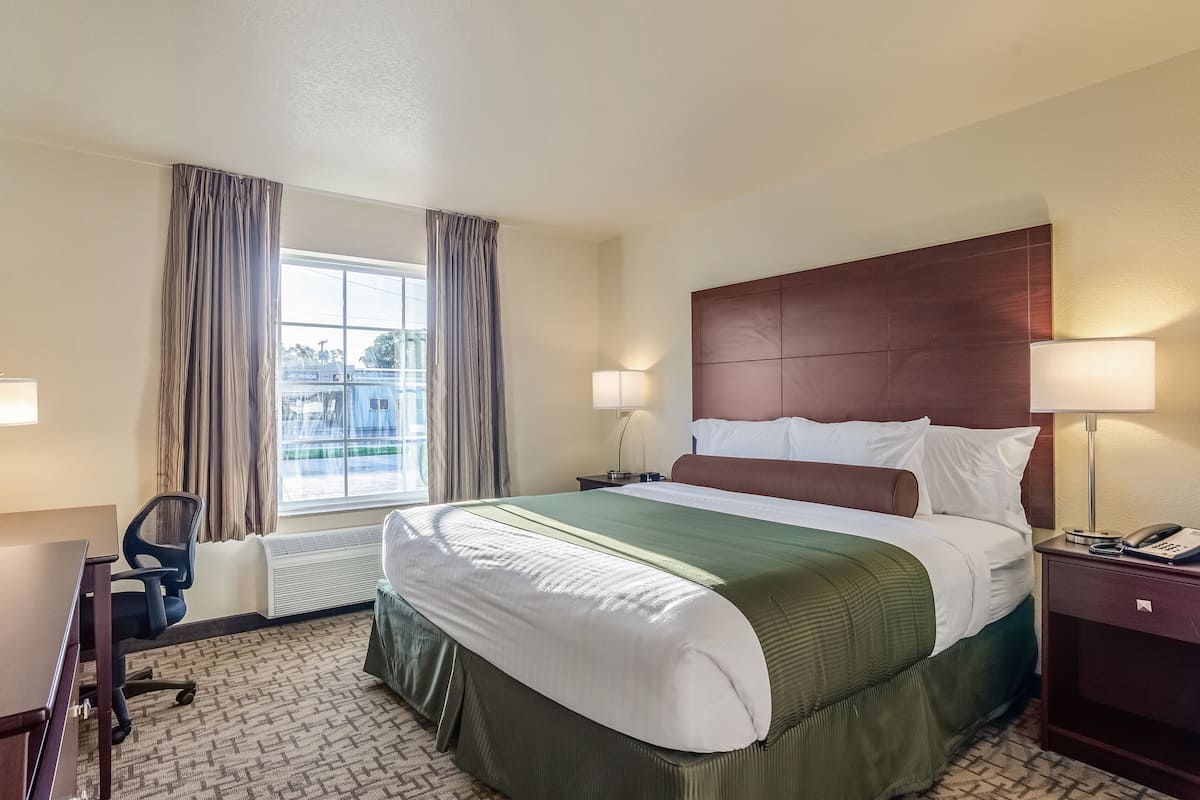 A hotel room with a large bed, two bedside lamps, a desk with a chair, and a window with curtains.