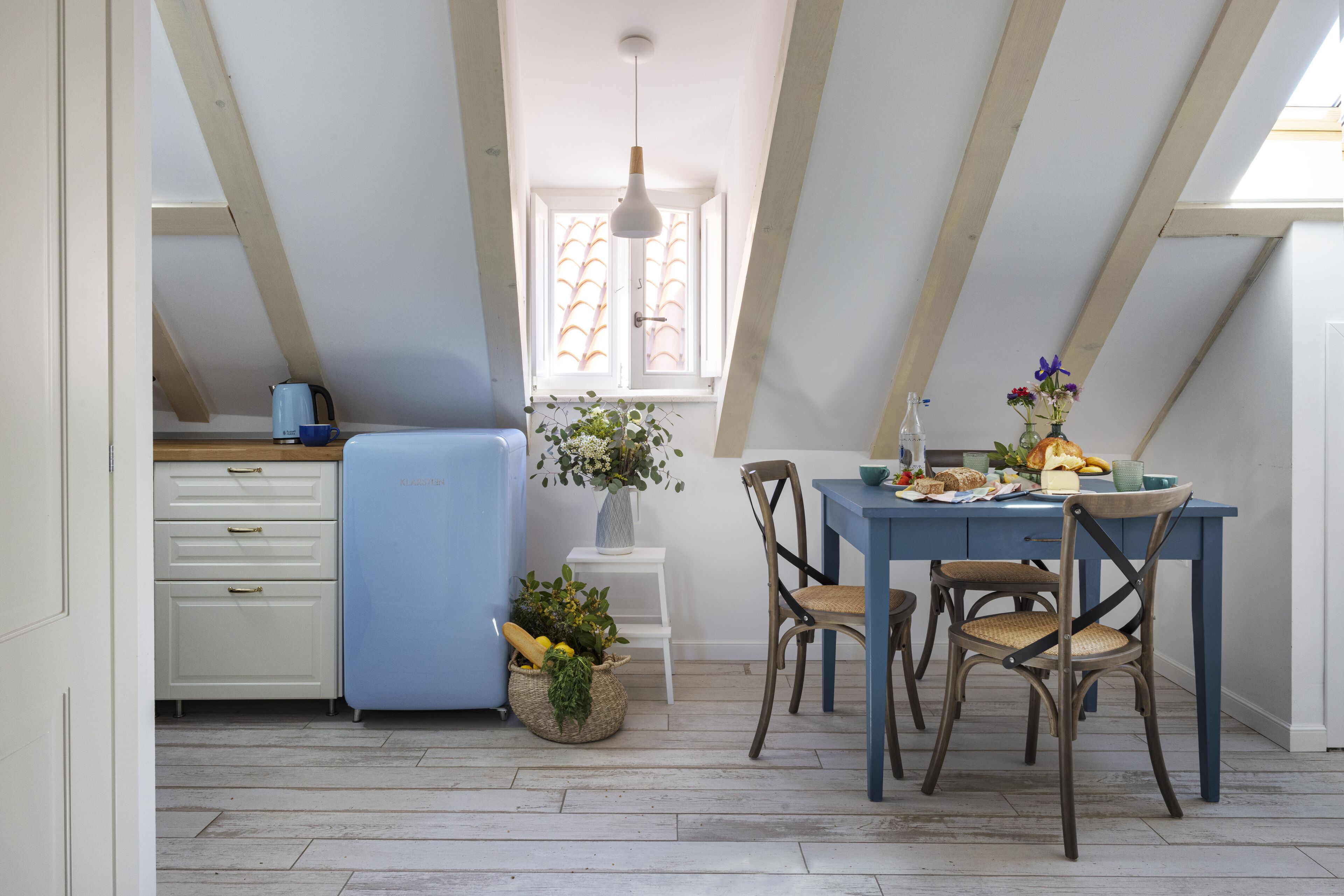 A kitchen with a blue refrigerator, wooden cabinets, a dining table with chairs, a window with curtains, and a hanging light fixture.