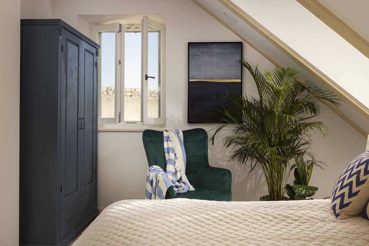 A bedroom with a bed, a green armchair, a potted plant, and a wardrobe.