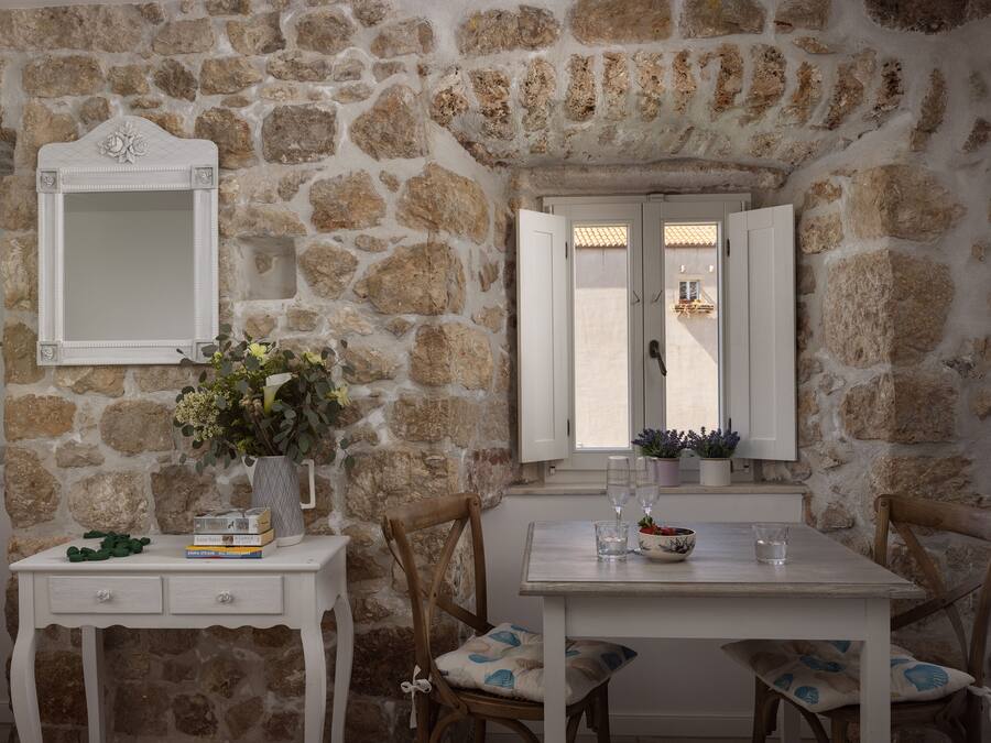 A stone-walled room with a white table and chairs, a mirror, and a window with shutters.