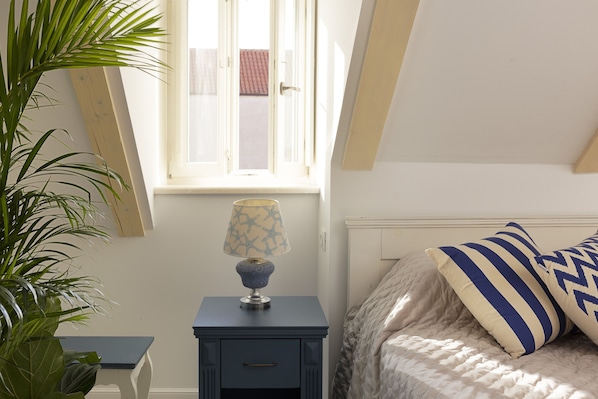 Un dormitorio con cama, una mesita de noche con lámpara, una ventana y una planta.