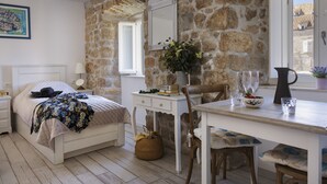 A bedroom with a stone wall, a bed with a patterned blanket, a bedside table with a lamp, a desk with a vase of flowers, and a window with a view of buildings.