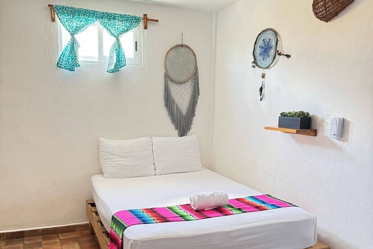 A bedroom with a wooden bed, white bedding, a colorful blanket, a ceiling fan, a dreamcatcher, a wall-mounted shelf with a plant, and a window with patterned curtains.