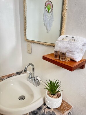 A bathroom with a white sink, a mirror with a rope frame, a shelf with toiletries, and a small plant. - Naay Bej (Tulum)