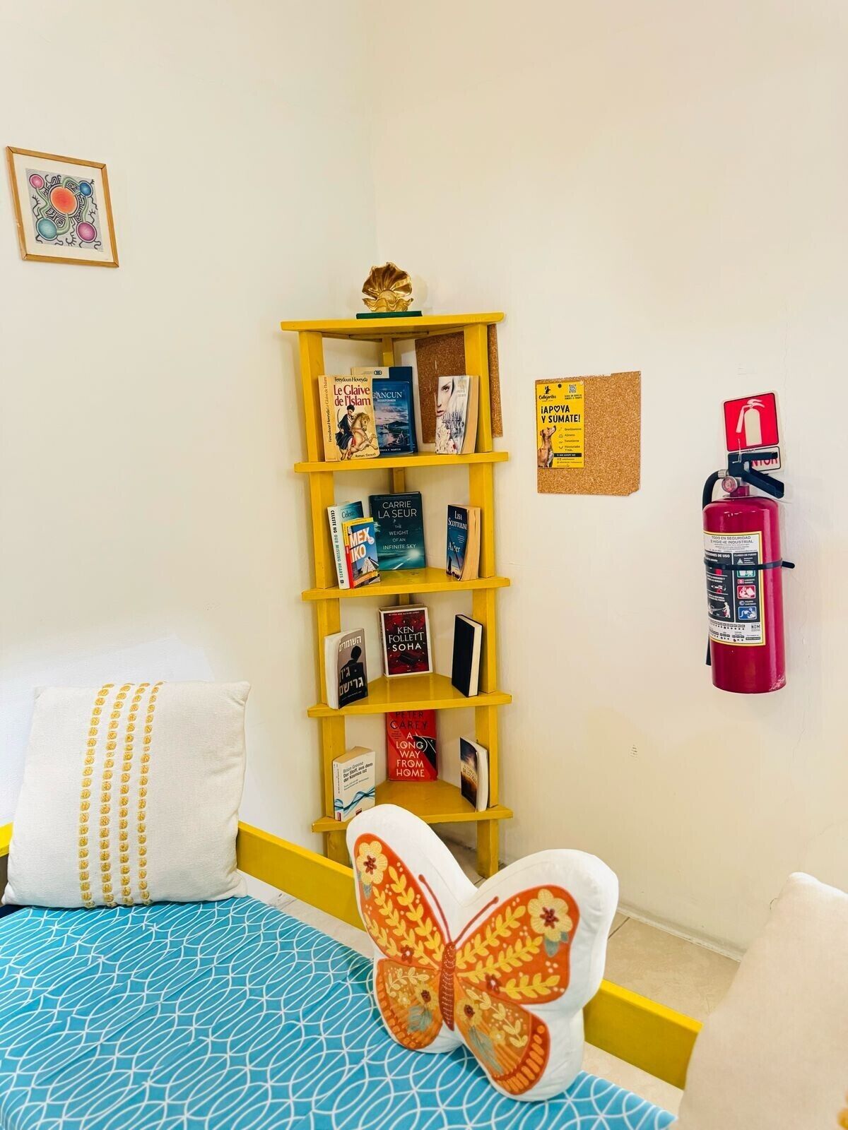 Una habitación con una cama, una estantería, un extintor y un tablón de anuncios.