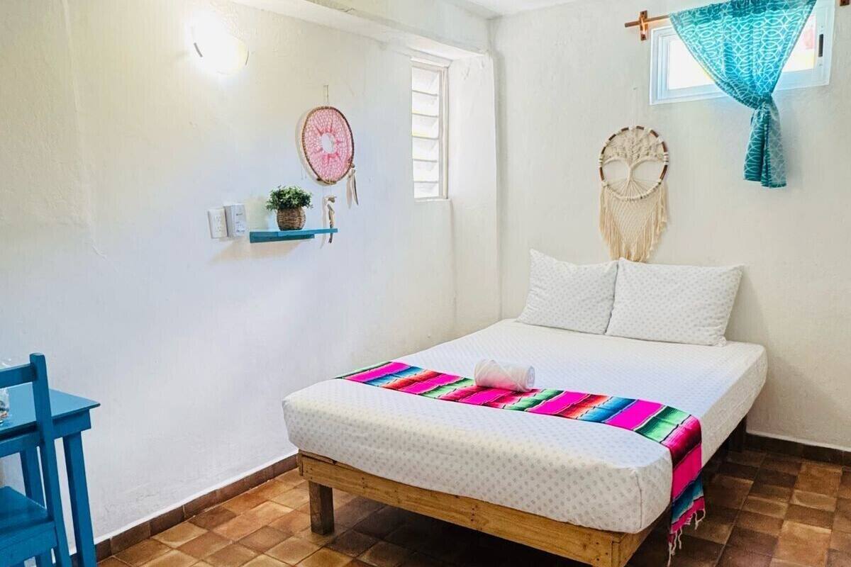 A bedroom with a bed, a ceiling fan, a window with a curtain, a shelf with decorative items, and a blue chair.