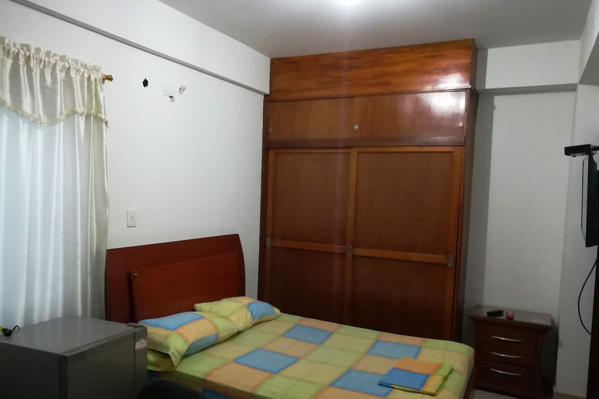 A bedroom with a wooden wardrobe, a bed with a colorful quilt, a desk, and a small bedside table.