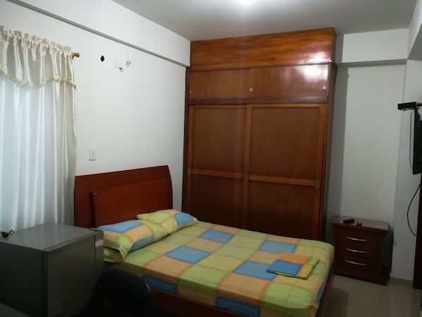 Un dormitorio con un armario de madera, una cama con colcha colorida, un escritorio y una mesita de noche.