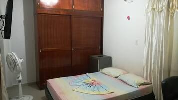 Un dormitorio con cama, armario, escritorio, silla, ventilador y cortina.