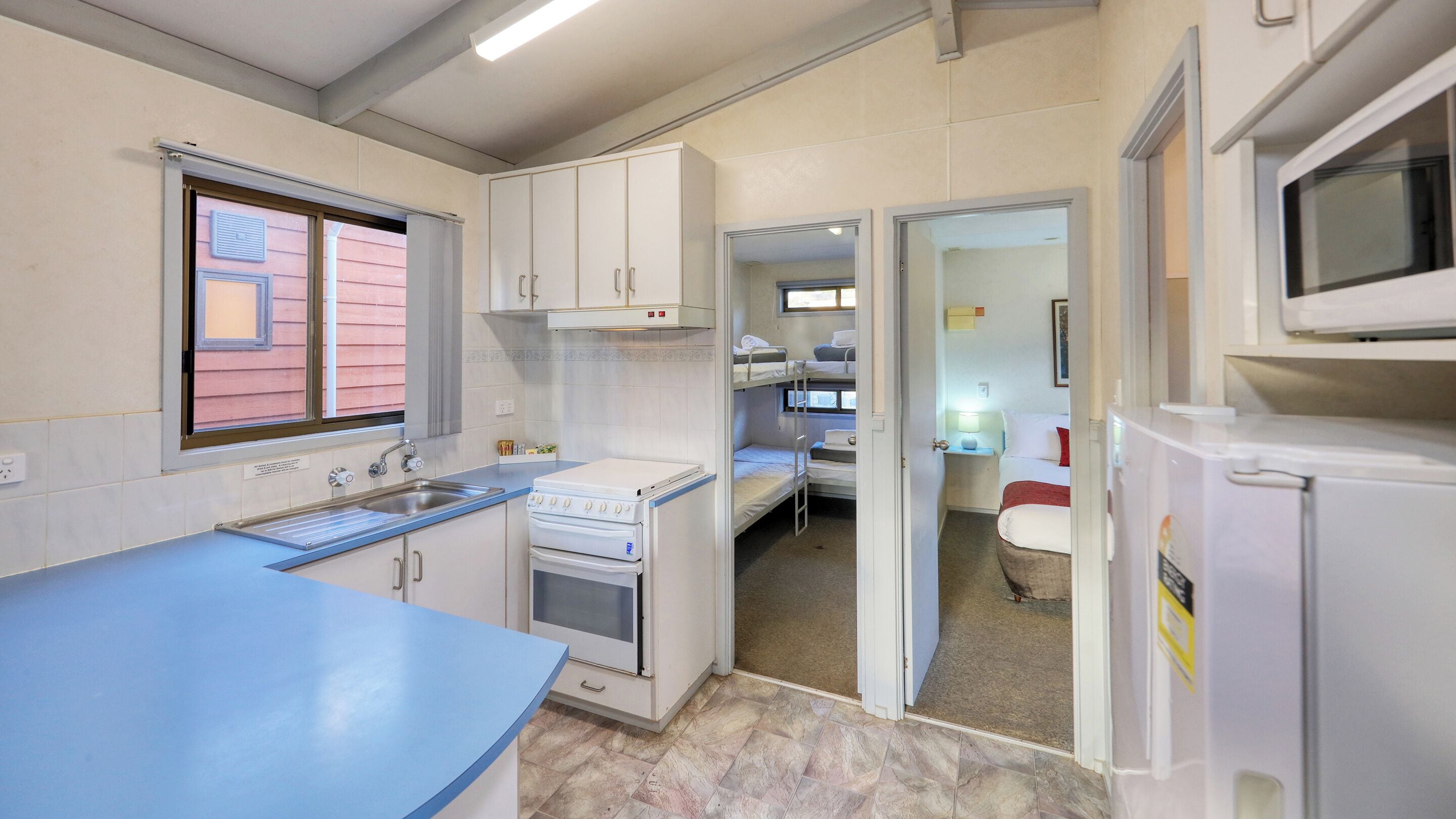 Una cocina compacta con un lavamanos, estufa y un refrigerador pequeño. Hay una ventana con persianas y una puerta que da a un dormitorio con literas.