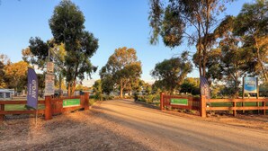 Une entrée fermée par une barrière donne accès au parc G'Day, avec des panneaux d'information, un chemin de terre et des arbres tout autour.
