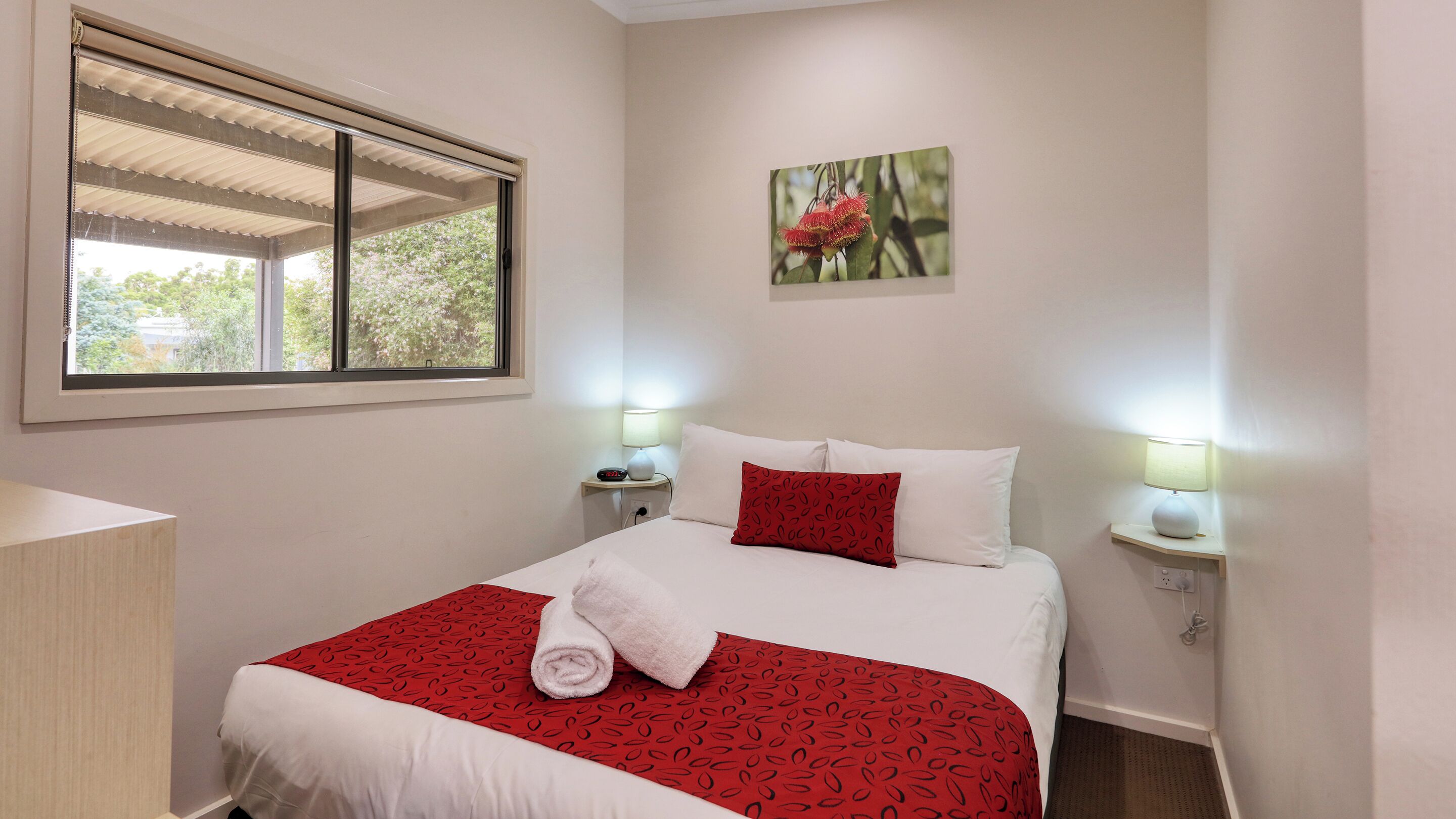 Cama bem arrumada com roupa de cama branca e vermelha, duas luminárias de cabeceira, um relógio de parede e uma janela com persianas.