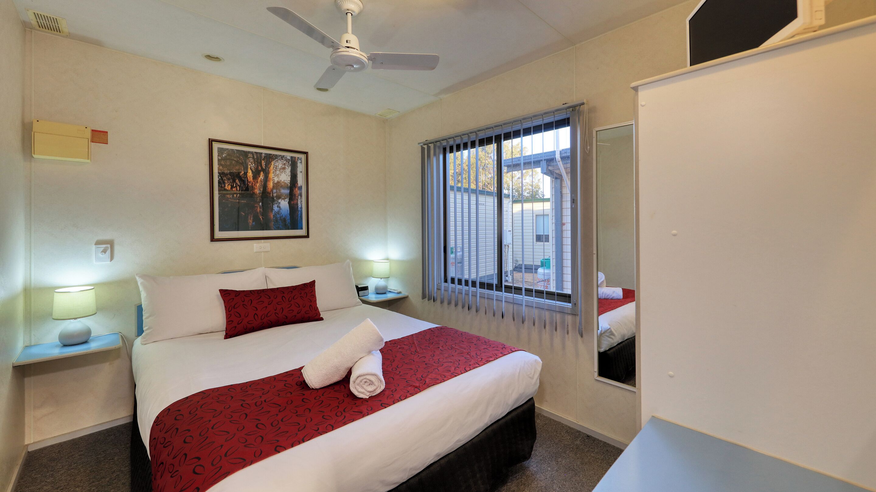 A hotel room with a bed, a nightstand, a lamp, a ceiling fan, a window with blinds, and a framed picture on the wall.