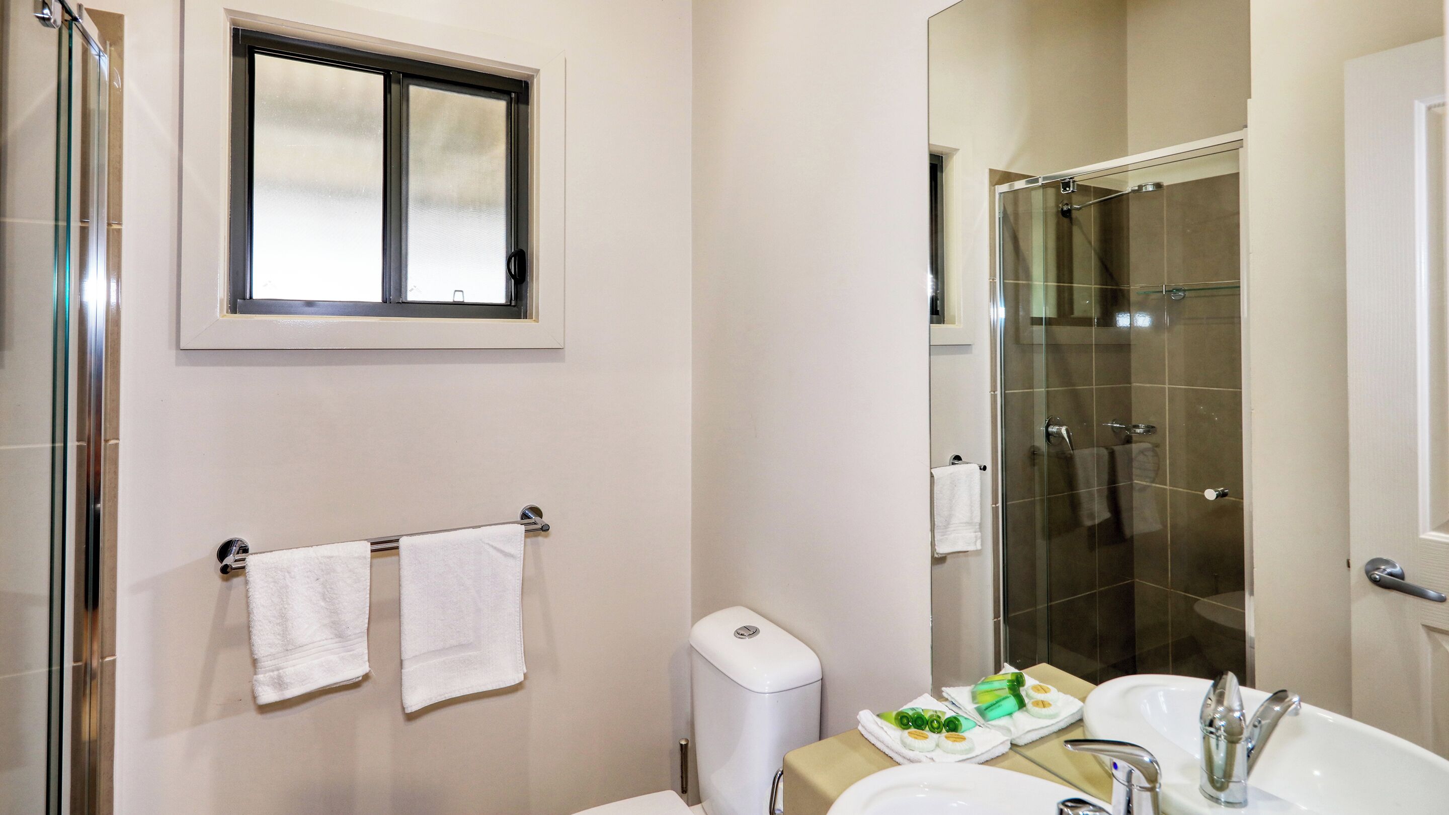 Un baño con cabina de ducha de vidrio, inodoro, lavabo con espejo y dos toallas colgadas en un perchero.