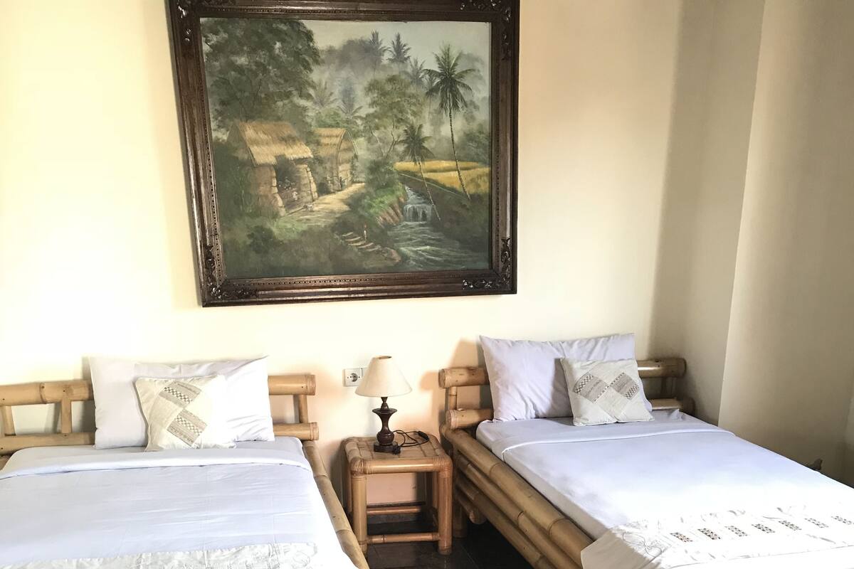 A room with two bamboo beds, a nightstand with a lamp, and a painting of a rural landscape on the wall.