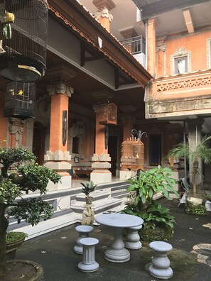 A courtyard with a traditional architectural style, featuring a central seating area with a round table and multiple stools, surrounded by potted plants and a birdcage. - Kacu guest house (Ubud)