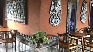 A room with a tiled floor, a wooden table and chairs, a potted plant, and decorative wall art. - Kacu guest house (Ubud)