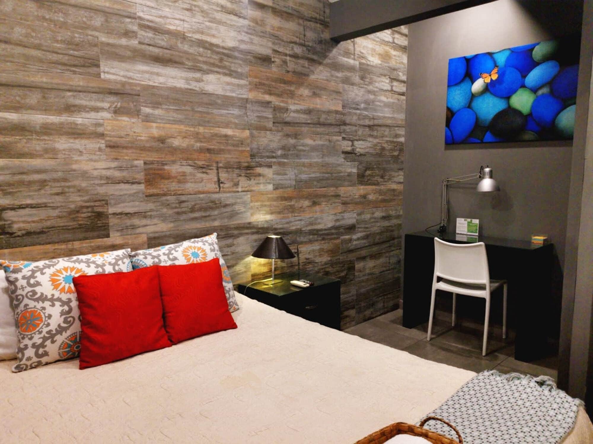A modern bedroom with a wooden wall, a bed with patterned pillows, a desk with a chair, and a painting of blue stones.