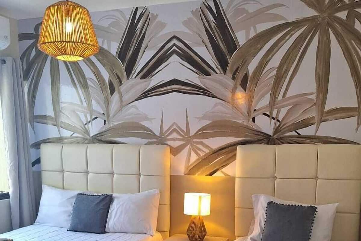 Ein Hotelzimmer mit zwei Betten, einem Nachttisch mit Lampe und einer Wandmalerei von Palmenblättern.