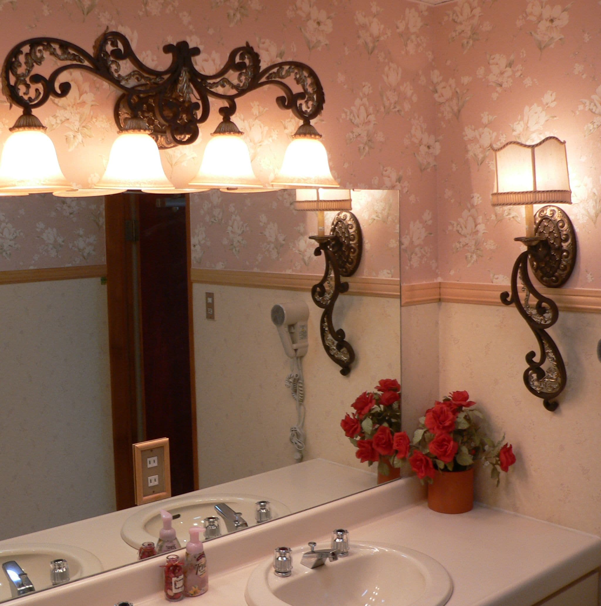 A bathroom with a double sink, a mirror with wall sconces, and a potted plant with red flowers.
