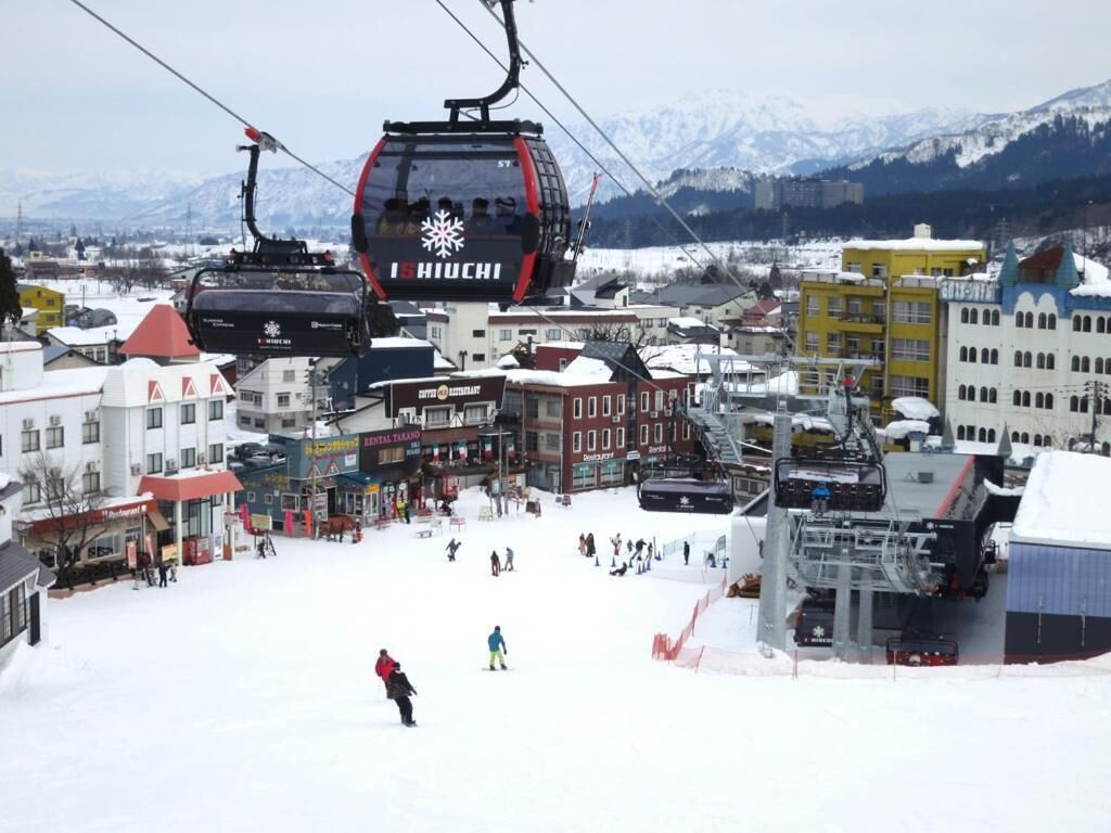 A ski lift with the name "Schiuchi" in a snowy town with buildings and mountains in the background.