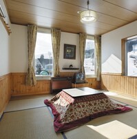Una habitación con paredes de madera y un ventanal grande. Se observa una cama con colcha estampada, una mesita pequeña y un televisor sobre un soporte de madera.