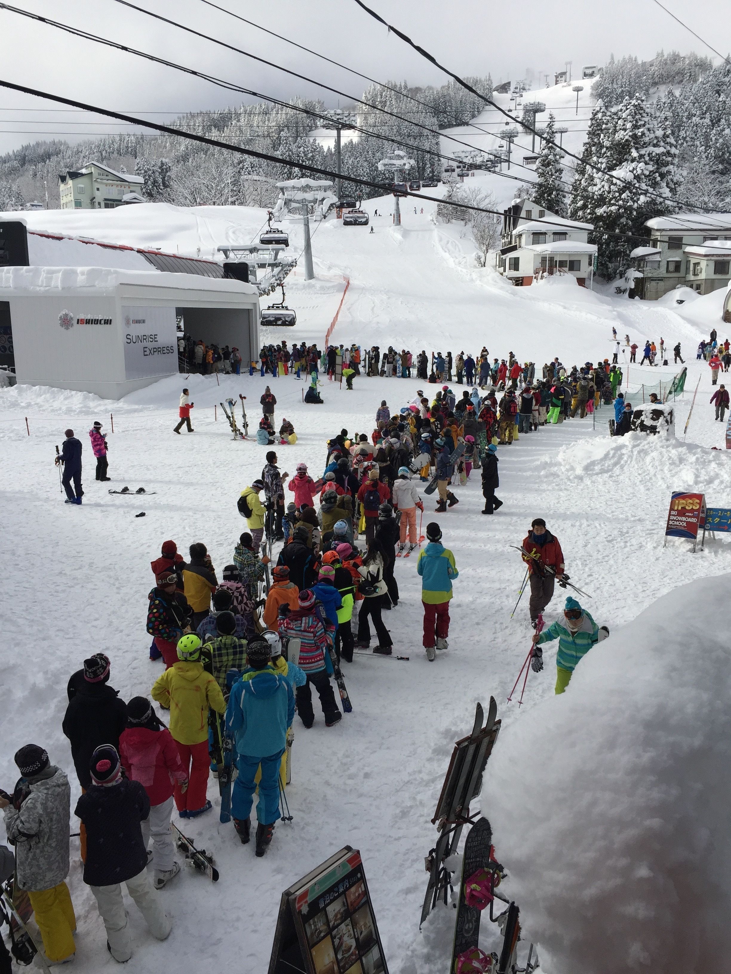 Une station de ski où une foule nombreuse s’est rassemblée près d’un télésiège, entourée d’arbres et de bâtiments recouverts de neige.