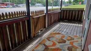 Eine Holzveranda mit einem gemusterten Teppich, einem weißen Zaun und Blick auf eine Straße und Grünflächen.