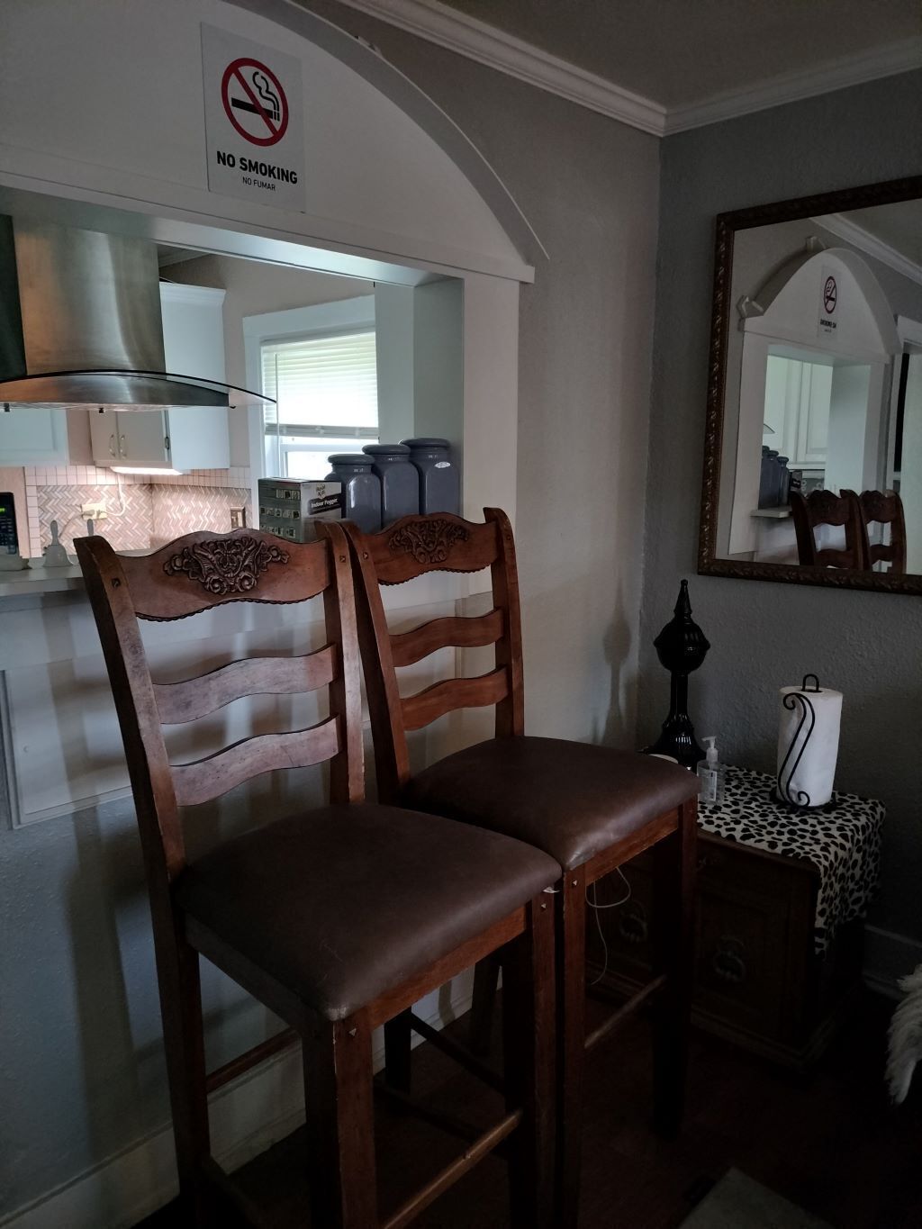 A dining area with two wooden chairs, a no smoking sign, a mirror, and a small table with a roll of paper towels.