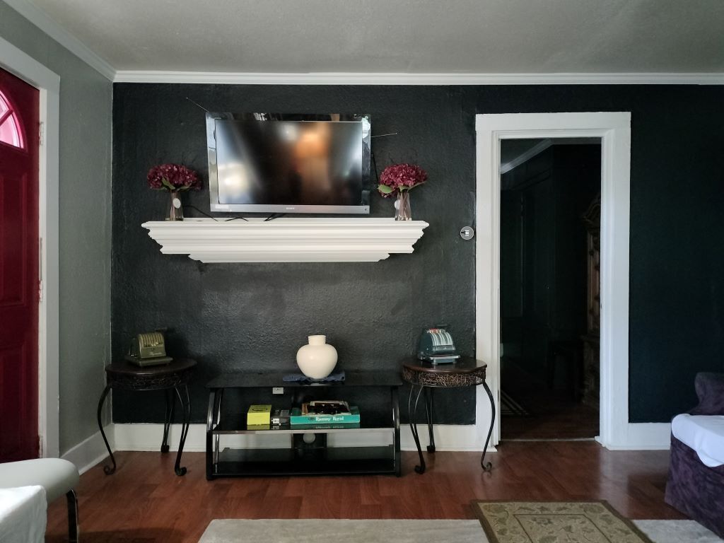 Un salon avec un téléviseur fixé sur un mur sombre, une cheminée blanche et une petite table ornée d’objets décoratifs.