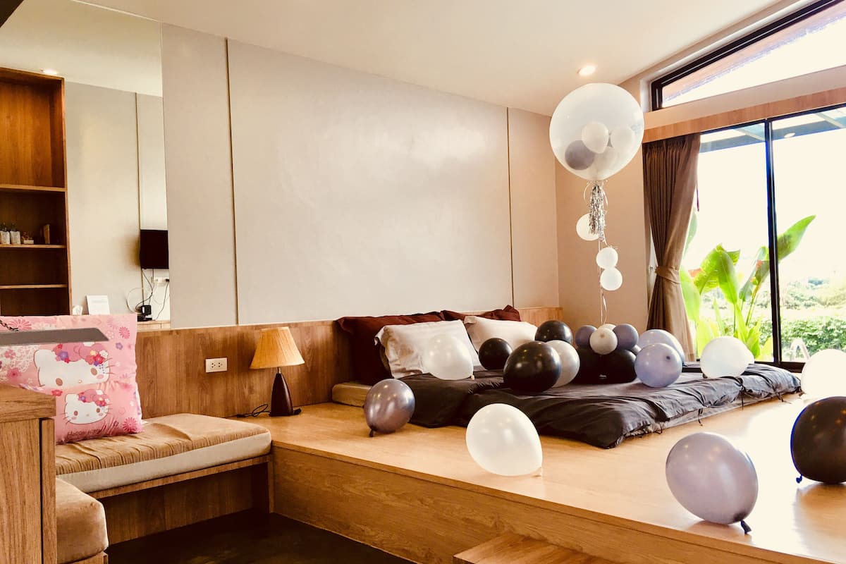 A modern bedroom with a large window, a wooden bed, and a seating area with a Hello Kitty cushion.