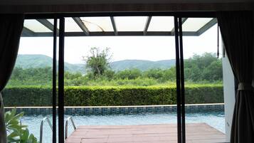 Habitación con un amplio ventanal corredero que da a una zona de piscina, vistas de exuberante vegetación y colinas, y una piscina con baldosas.