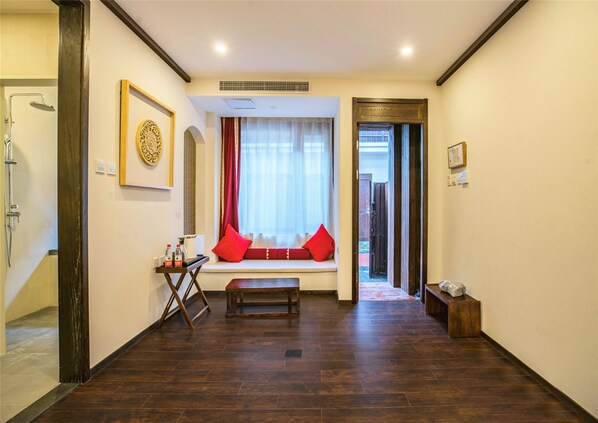 Una habitación de hotel con piso de madera, un banco, una ventana con cortinas rojas y una puerta que da a un espacio al aire libre.