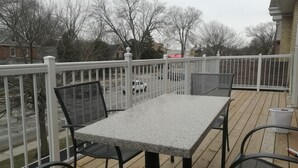 A covered outdoor deck with a table and chairs, a white railing, and a view of a street with cars and trees. - Ambassador's Inn Next Door (Stratford)