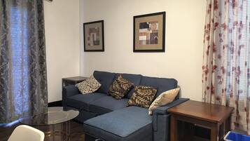 A living room with a blue sofa, two framed abstract paintings, a glass coffee table, a wooden side table, and a blue trash bin.