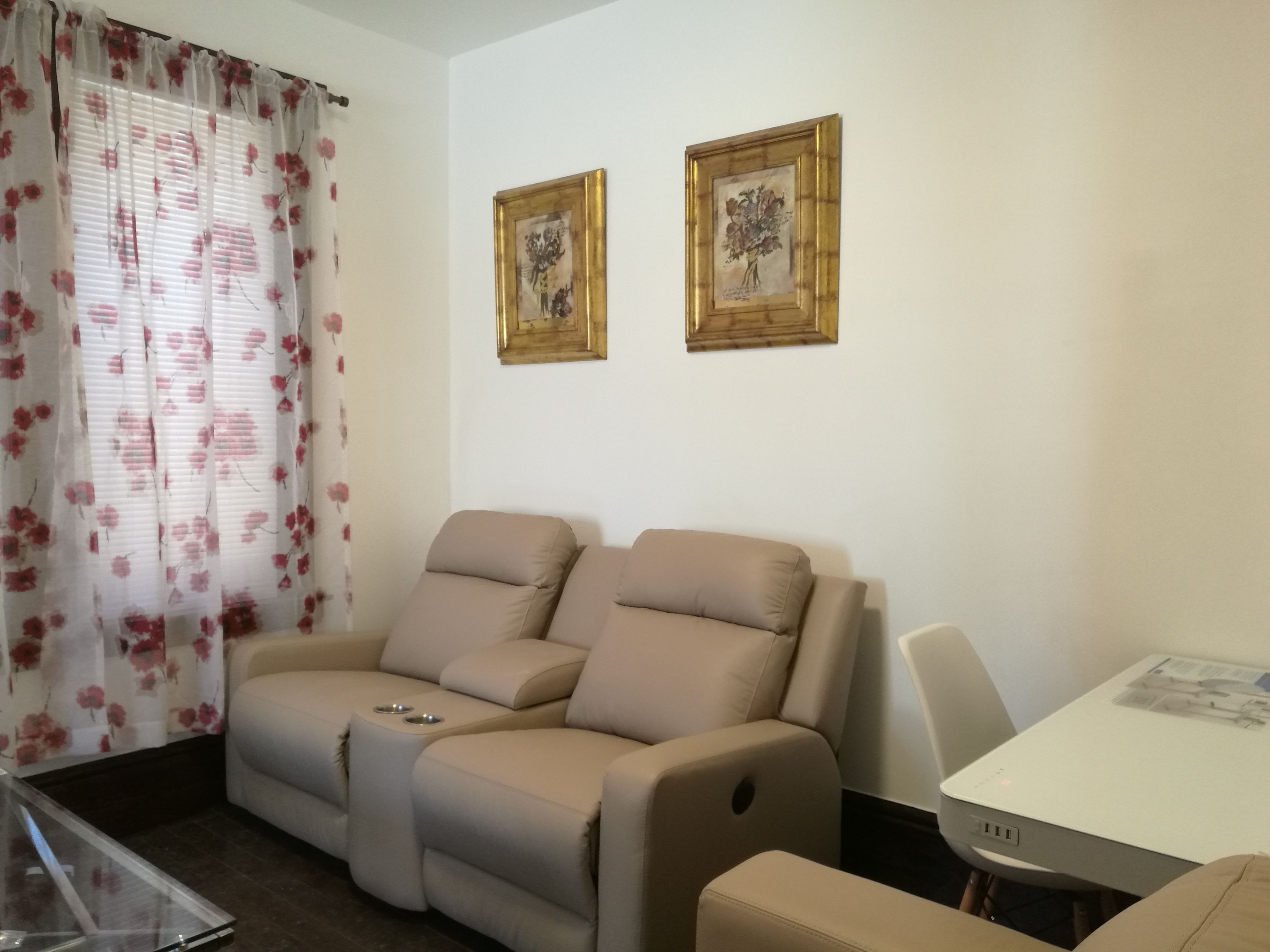 A living room with a beige sofa, a glass coffee table, and a white dining table. There are two framed botanical prints on the wall.