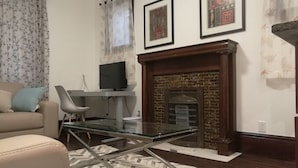 A living room with a fireplace, a sofa, a glass table, and a TV on a stand. - Ambassador's Inn Next Door (Stratford)