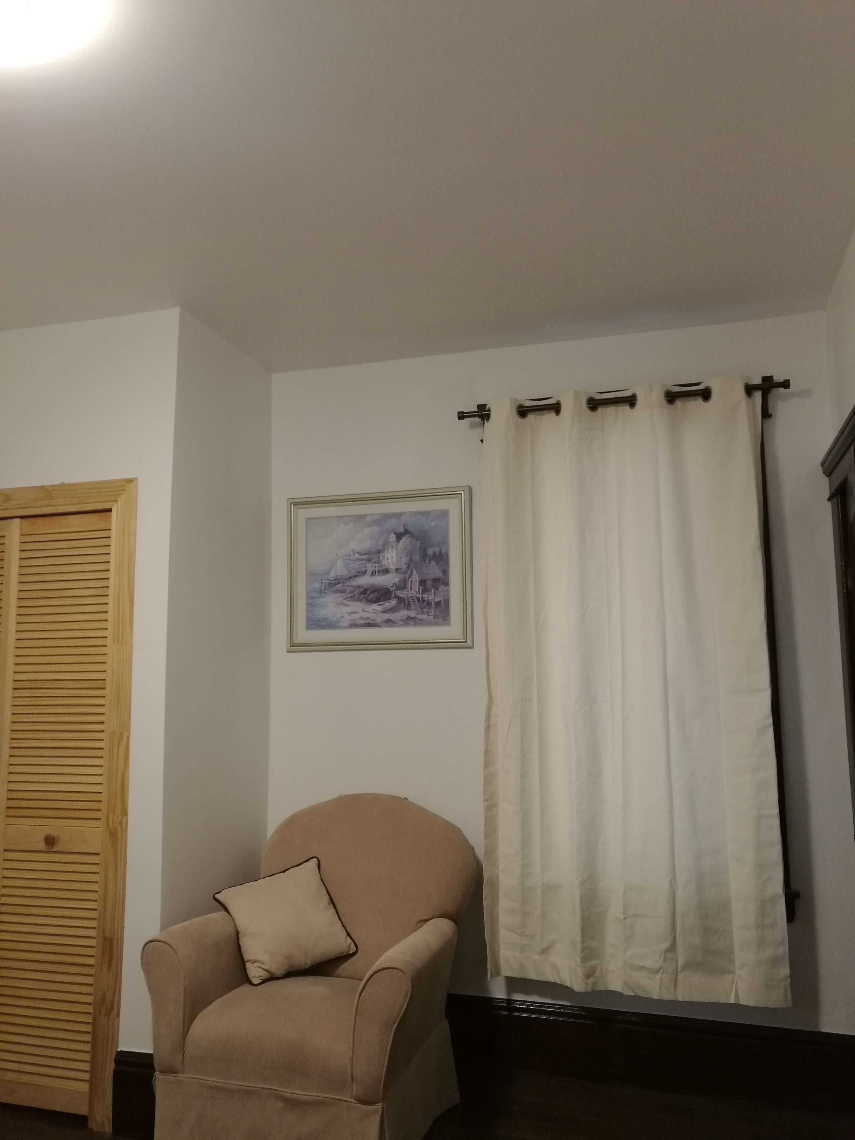 Une pièce avec un fauteuil beige, une porte en bois, un tableau encadré au mur et un rideau blanc.
