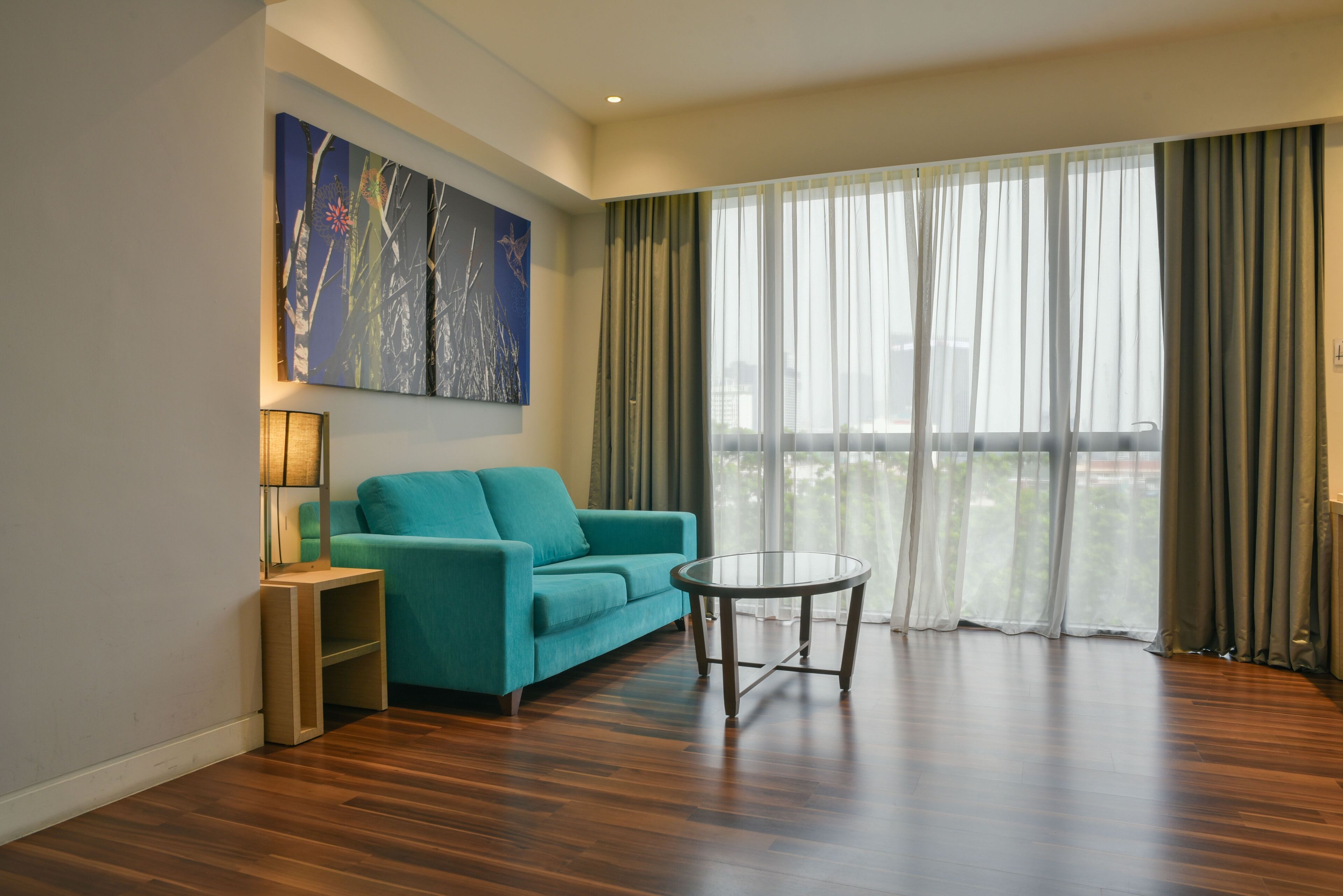 Una sala de estar moderna con un sofá color turquesa, una mesa de centro de vidrio y un ventanal con cortinas de encaje.