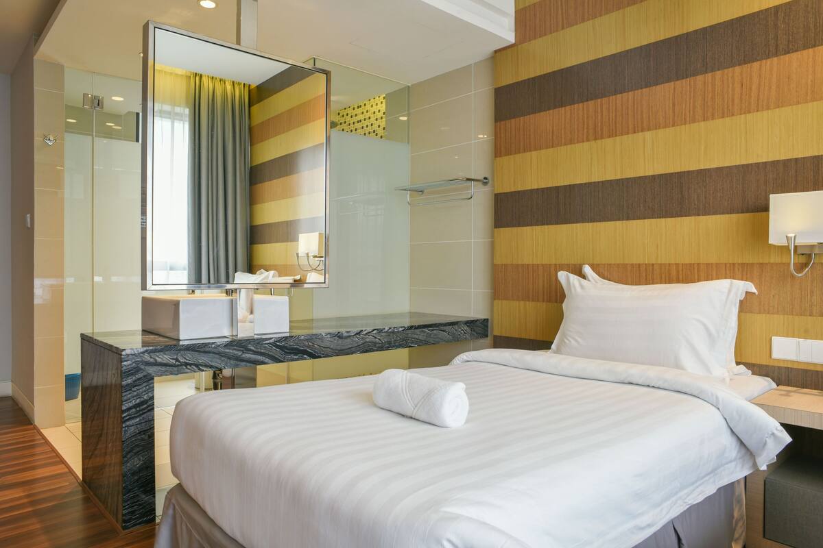 A modern hotel room with a large bed, a glass shower enclosure, a marble countertop, and a striped wall design.