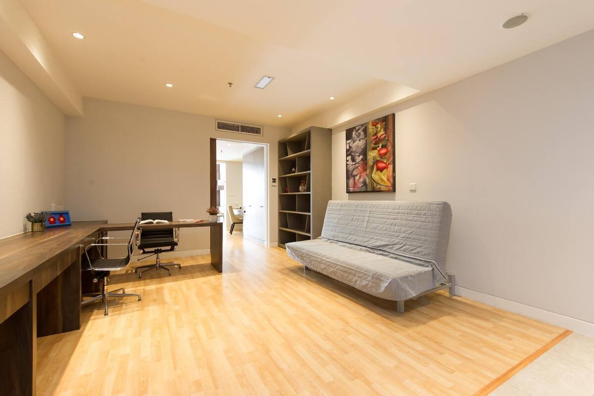 A modern room with a wooden floor, a desk with a chair, a grey sofa, and a bookshelf.