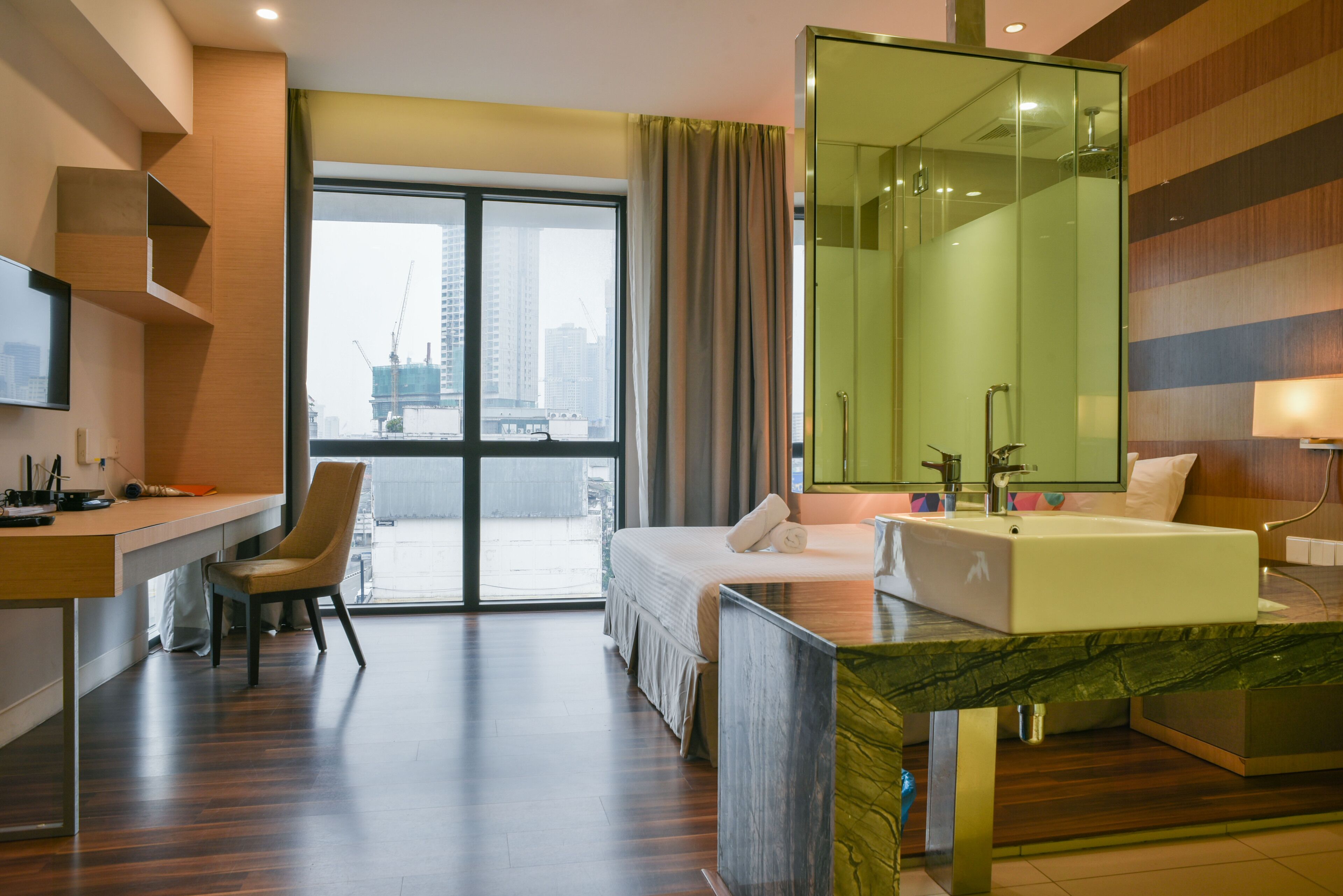 A hotel room with a bed, a desk, a chair, a large mirror, and a view of the city through the window.