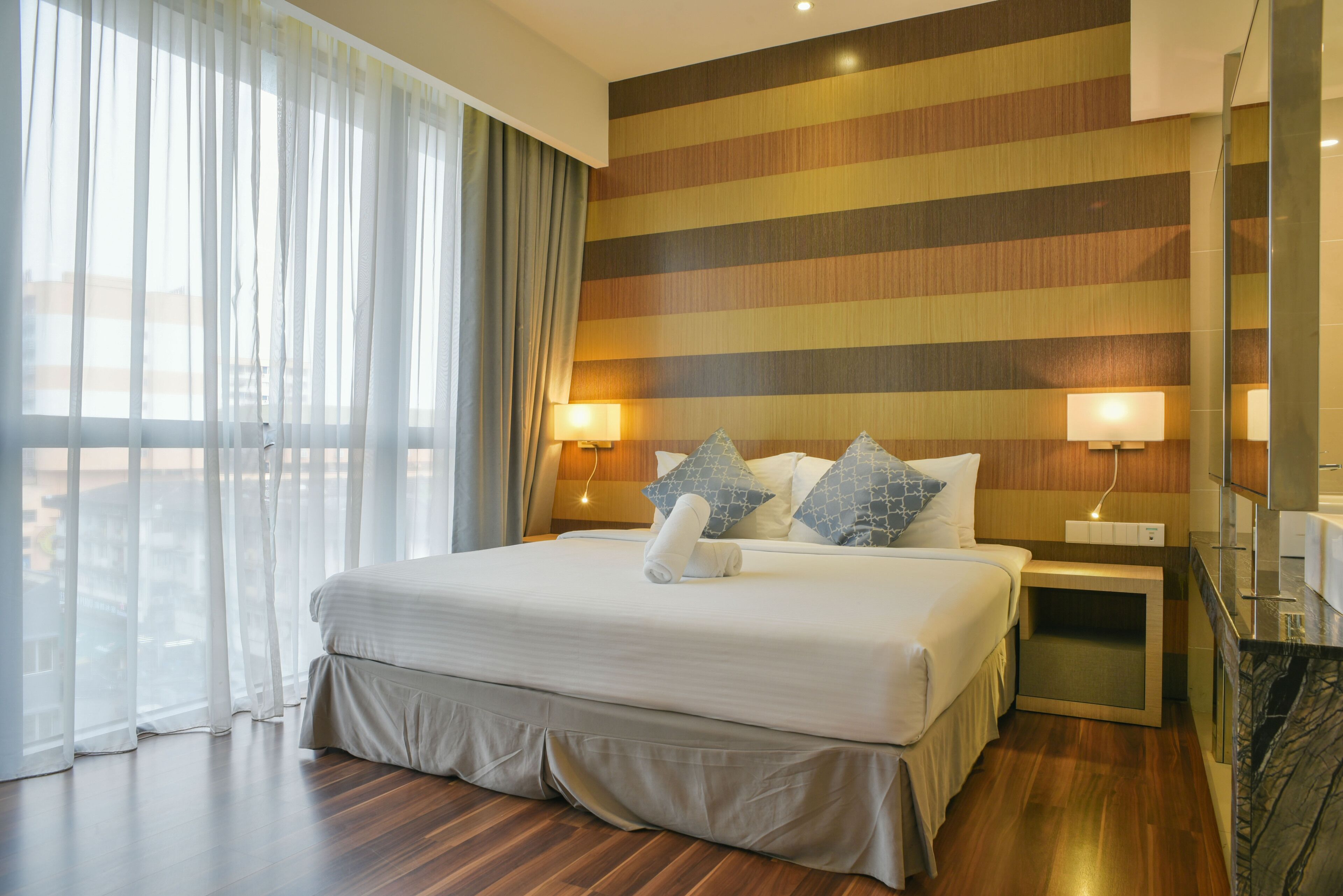 Quarto de hotel com uma cama grande, duas luminárias de cabeceira, um painel de cabeceira de madeira e vista através de cortinas leves.