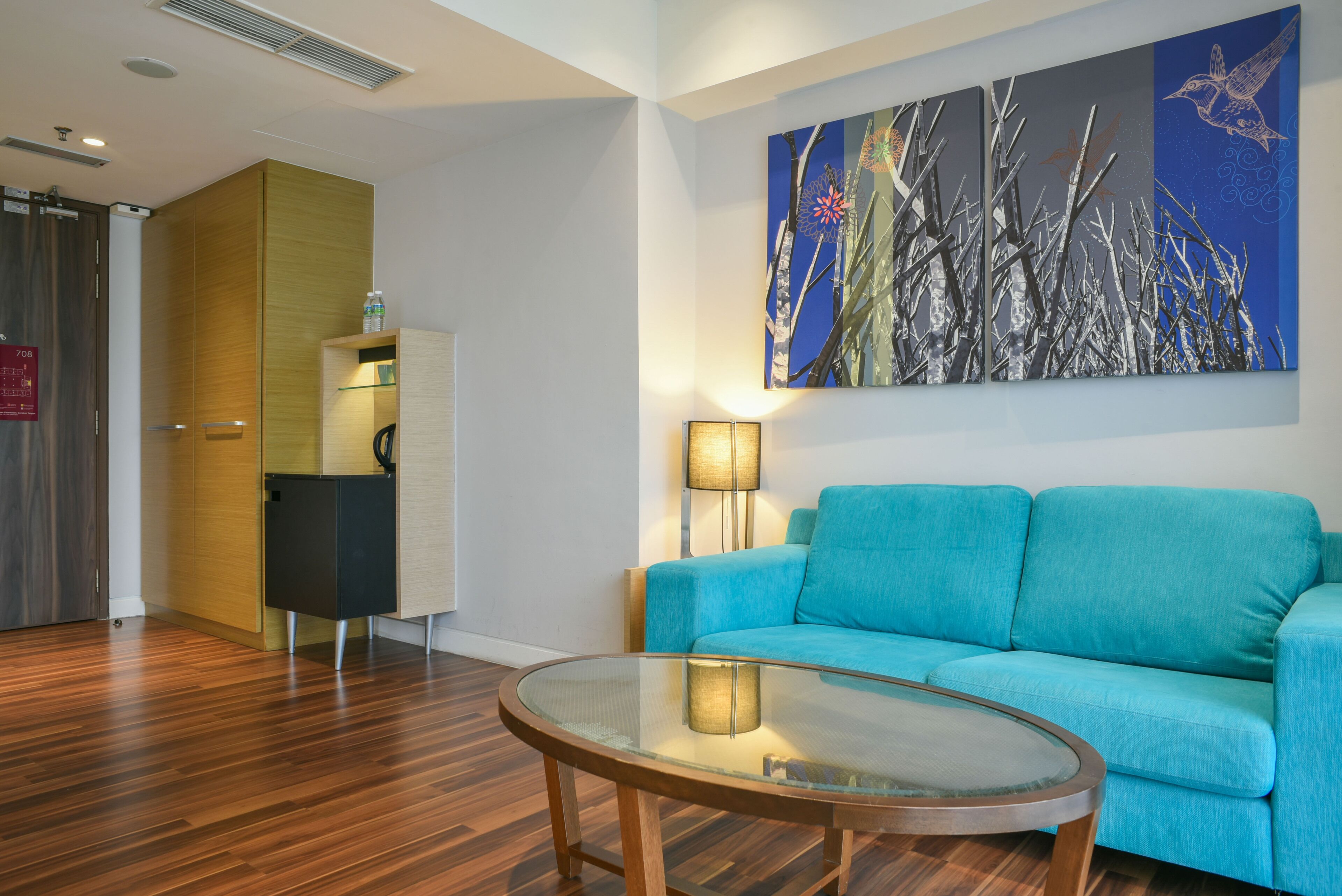 A modern living room with a blue sofa, a glass coffee table, and a painting of trees on the wall.